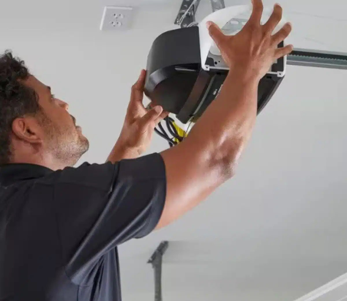 A man adjusts or installs a garage door opener on the ceiling, using both hands to secure the device. He is wearing a black shirt and looking up at the equipment while working.