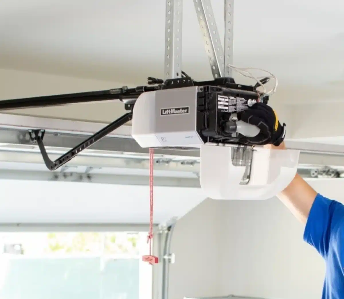 A person’s arm adjusts a LiftMaster garage door opener mounted on the ceiling, with a red pull cord hanging down and the garage door partially open in the background.