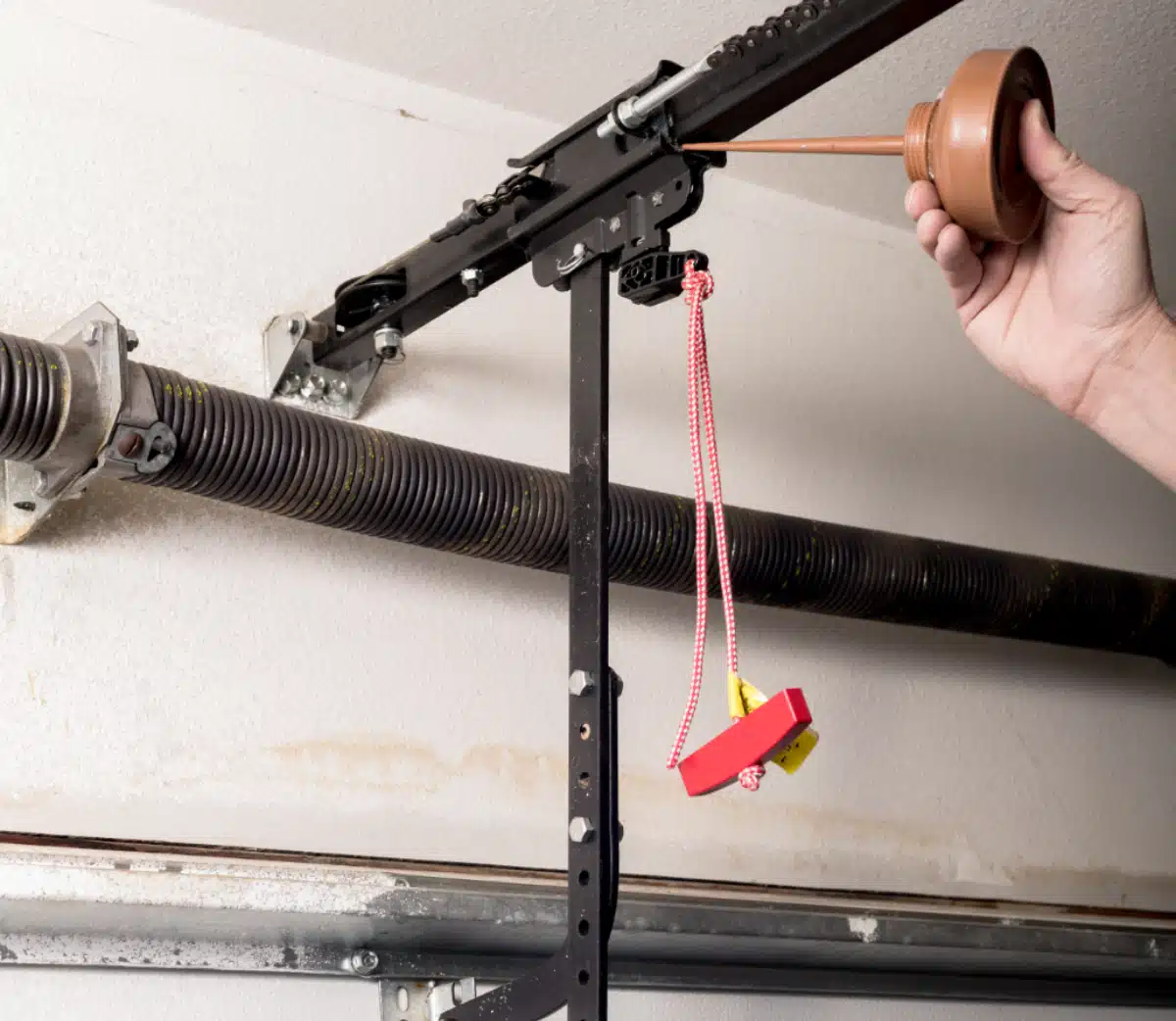A hand applies lubricant oil to the chain of a garage door opener mechanism mounted on the ceiling; a red emergency release cord hangs from the opener.