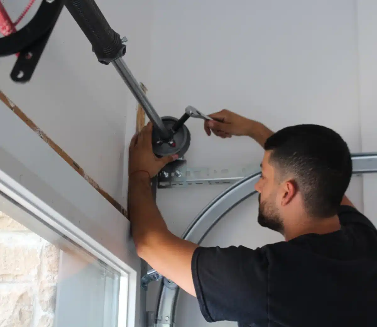 A man uses a wrench to adjust a garage door torsion spring mounted on the wall above a window, working on the door mechanism.