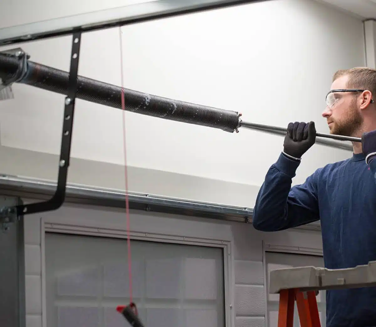 A man wearing safety glasses and gloves stands on a ladder, repairing or installing a garage door torsion spring using a metal rod inside a well-lit garage.