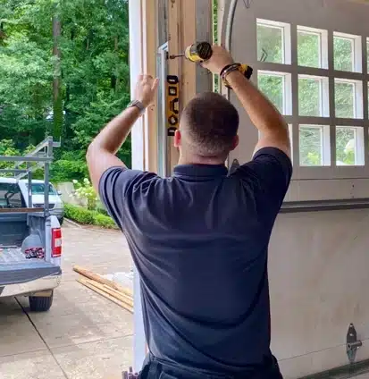 A person in a dark polo shirt uses a drill and a level to install or repair a garage door opener rail inside a garage, with a pickup truck parked outside in the driveway.
