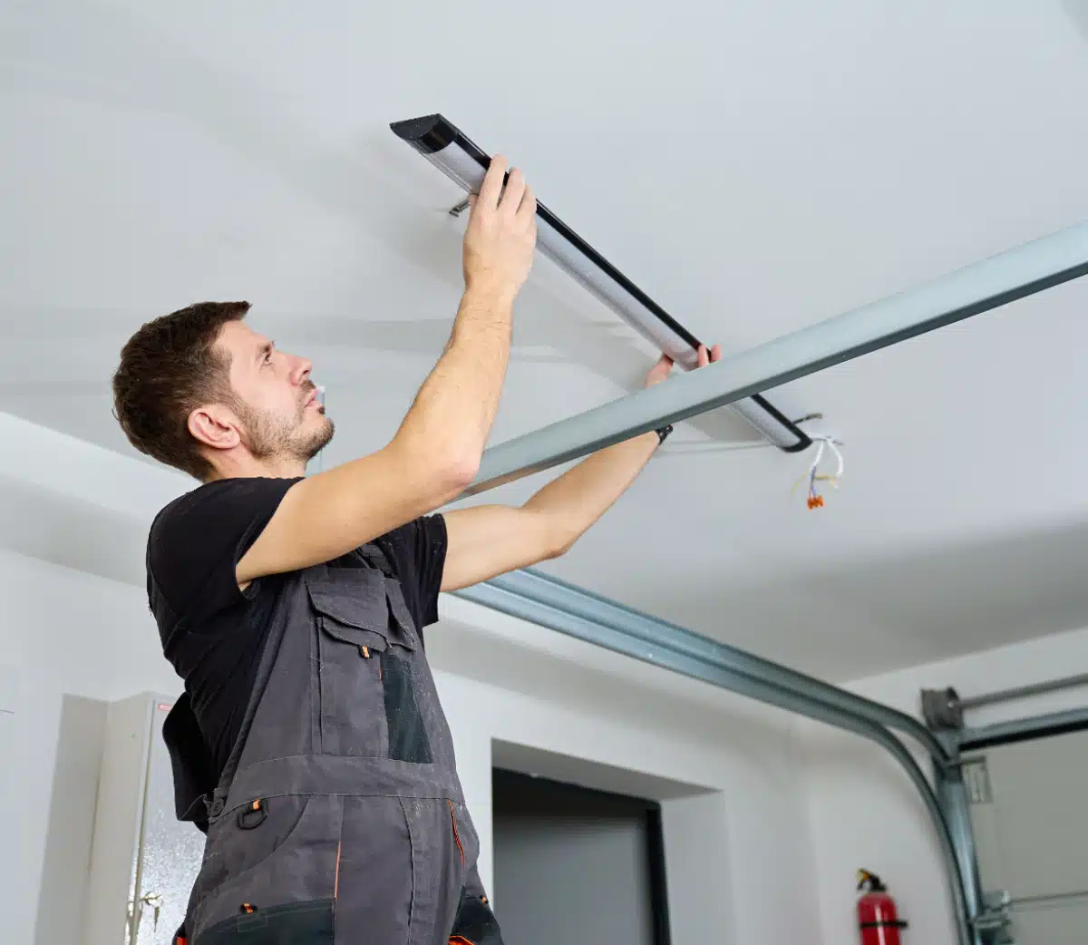 A man in work overalls stands on a ladder and installs or repairs an automatic garage door opener on the ceiling of a garage. He is using tools and focusing on attaching a metal rail.