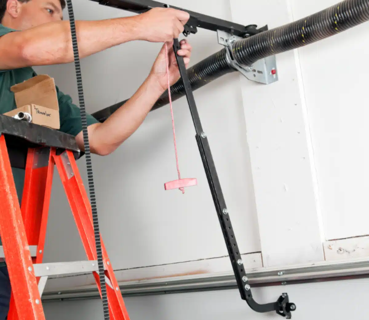 A person on a red ladder repairs a garage door mechanism, holding a metal bar with an emergency release cord and handle near the garage ceiling.