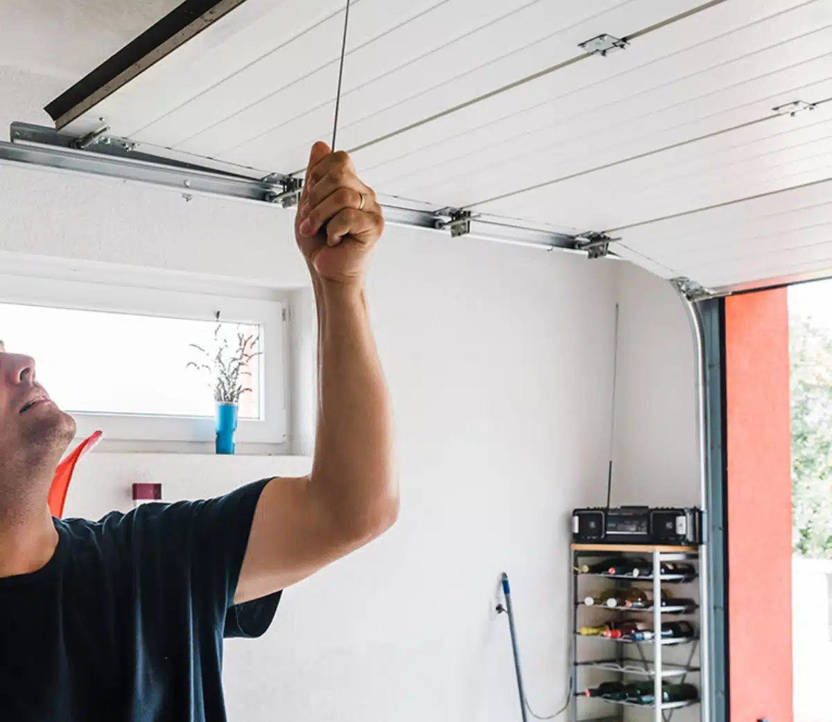 A person in a garage pulls an emergency release cord to manually open a white garage door. Shelves with various items and a window are visible in the background.